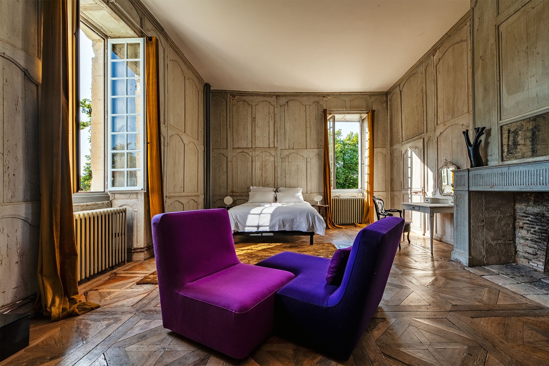 A panelled château bedroom with tall windows, a fireplace and coloured armchairs